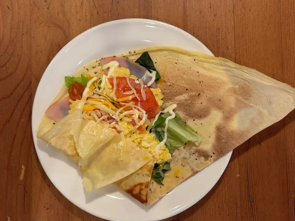 お食事用クレープ。ハム、スクランブルエッグ、チーズ、レタス、トマト、もやしを包んだ四角い盛り付けの一皿、木製テーブルに白い皿で提供