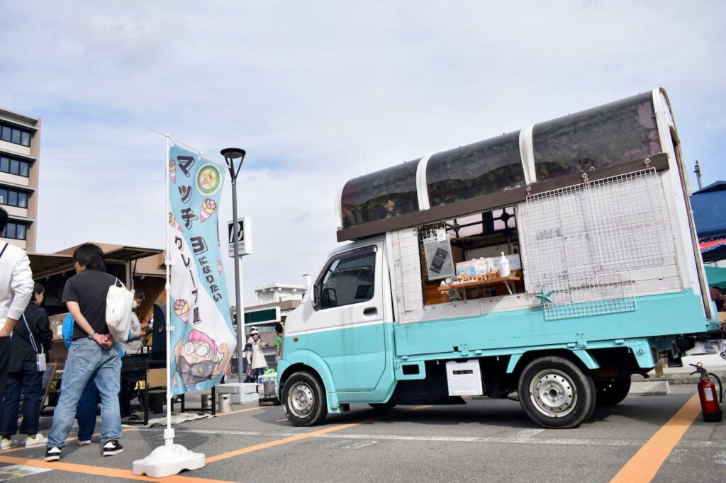 クレープ移動販売車「パライソクレープ」。水色と白のツートンカラーのキッチンカーで、屋根には大きな茶色の看板が設置されている。車の横には青いのぼり旗が立っており、クレープのイラストが描かれている。屋外イベント会場での営業風景。