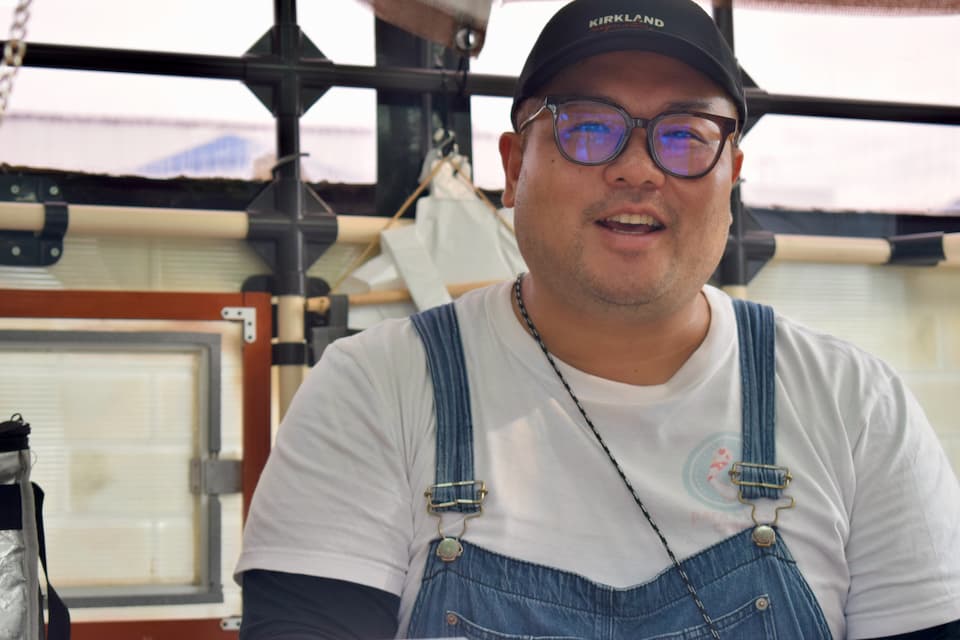 黒いキャップと眼鏡をかけた パライソ・クレープのシェフ水野氏が、白いTシャツにデニムのエプロンを着用してキッチンカー内で笑顔を見せている様子