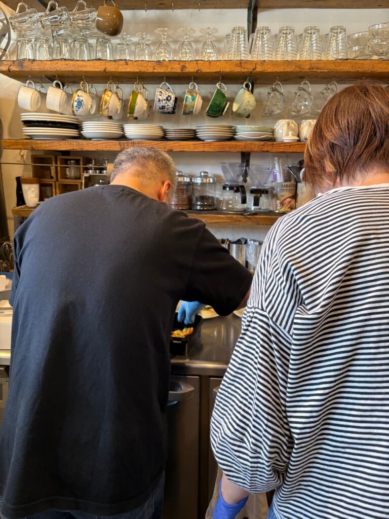 木製棚に色とりどりのティーカップやグラス、皿が並ぶカフェのカウンター。青いゴム手袋をつけた黒いトップスの人物と、白黒ボーダーのトップスの人物が、ステンレスカウンターで料理を盛り付けている。背後の棚にはコーヒー豆の瓶や器具が並ぶ。
