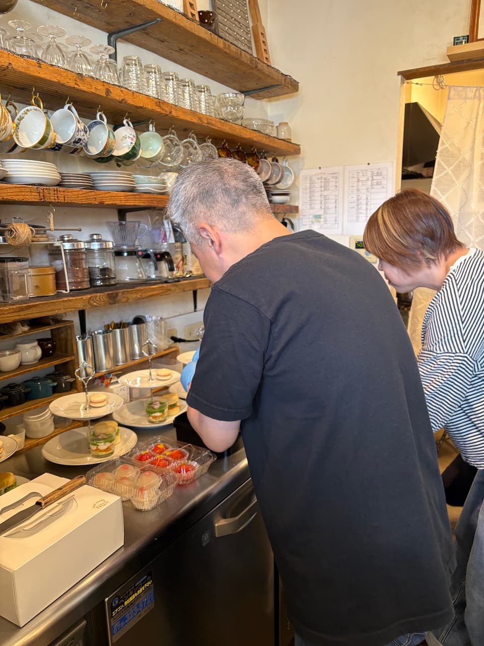 木製棚にティーカップやグラス、調理器具が並ぶカフェのカウンター。黒いTシャツに青いゴム手袋の人物と白黒ボーダーの人物が、ステンレスカウンターでアフタヌーンティースタンドを組み立てている。手前にはいちごなどのフルーツパックと白い箱が置かれている。