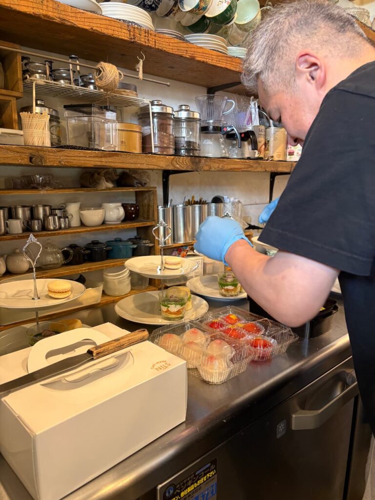 
青いゴム手袋をつけた黒いTシャツのスタッフが、道具や食器が並ぶ木製棚前のステンレスカウンターで、2段スタンドにスイーツを盛り付けている。手前にはケーキボックス、いちごのフルーツパックが並び、左奥にはマカロンをのせた別のスタンドも見える。